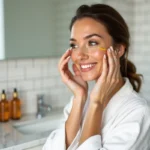 Femme appliquant un sérum doré sur son visage dans une salle de bain minimaliste, comparaison soins de peau Caudalie et Nuxe