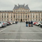 Zone de stationnement gratuit devant le château de Versailles avec des voitures garées et des touristes marchant vers l'entrée